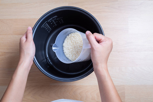 Το γυναικείο χέρι ρίχνει ρύζι γιασεμί στην κατσαρόλα. Χρησιμοποιώντας γυαλί μέτρησης. σε ξύλινο τραπέζι. - Φωτογραφία, εικόνα