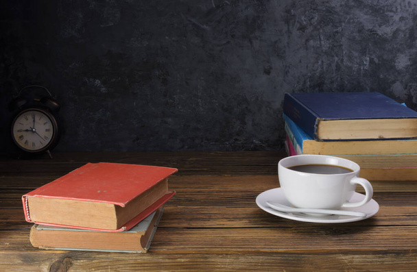Λευκό φλιτζάνι καφέ με σκούρο μαύρο καφέ και παλιά βιβλία Και το ρολόι είναι τοποθετημένα σε ένα παλιό ξύλινο τραπέζι και ένα μαύρο ξύλινο τοίχο. - Φωτογραφία, εικόνα