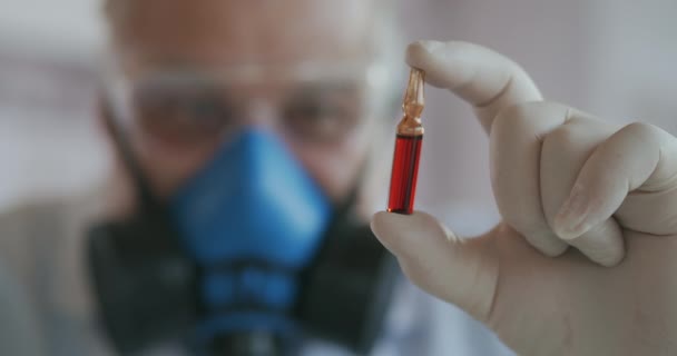 Extrême Gros plan d'un scientifique dans un respirateur bleu et des lunettes de protection, développeur d'un vaccin contre le coronavirus tenant une ampoule rouge. Le médecin examine le nouveau vaccin. Un nouveau type de virus  - Séquence, vidéo