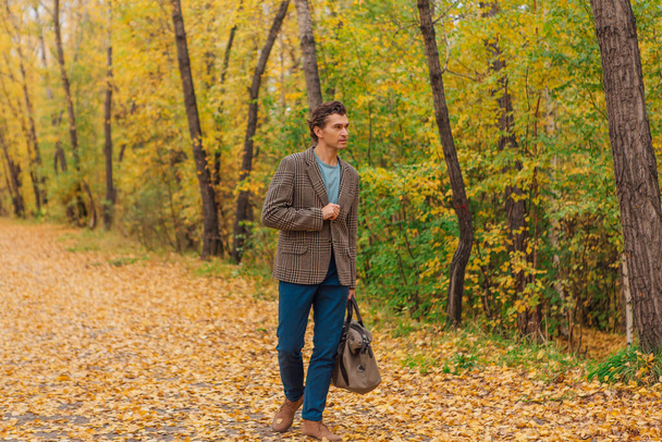 Высокий красивый мужчина в коричневой куртке ходит с сумкой на осеннем переулке - Фото, изображение