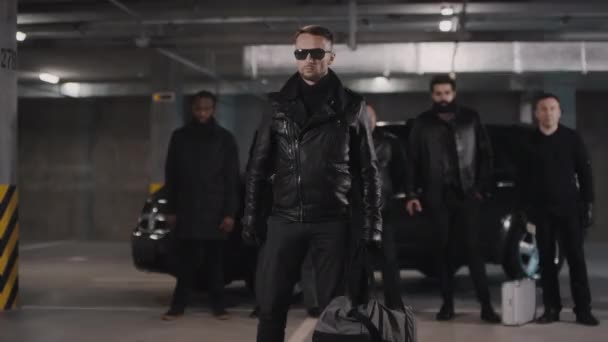 Средние кадры, где мужчина-кавказский преступник несет темную спортивную сумку с деньгами, снимающими черные солнцезащитные очки, стоящие на изолированной подземной парковке впереди своей преступной банды - Кадры, видео