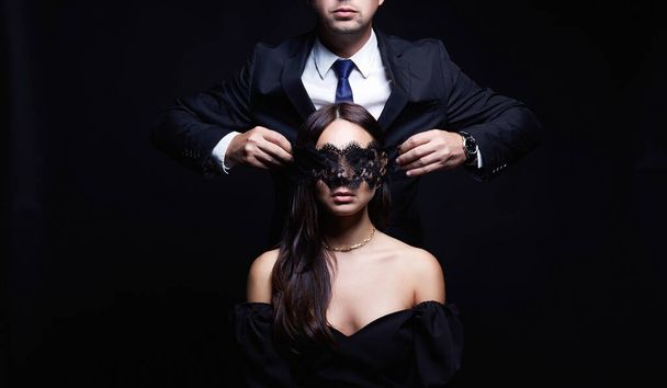 красивая женщина в маске и мужчина в костюме. Пара на черном фоне. Очаровательная, элегантная девушка соблазняет своего красивого парня - Фото, изображение