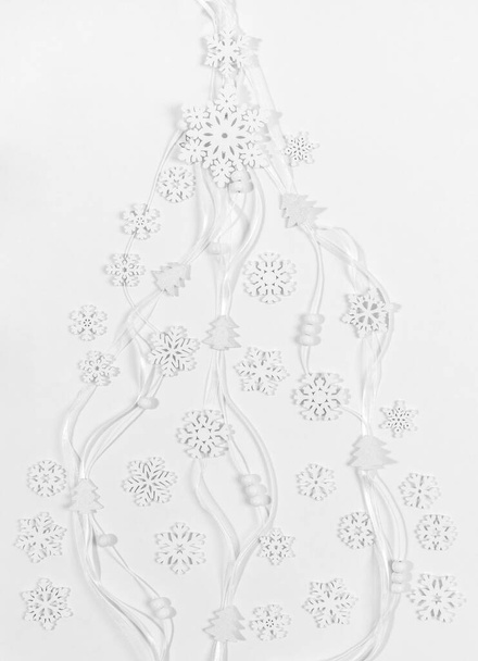 Festive trendy Christmas white background. White wooden snowflakes and small Christmas trees and round beads on narrow ribbons laid out as creative Xmas tree. Xmas greeting card. Top view, flat lay. - Fotoğraf, Görsel