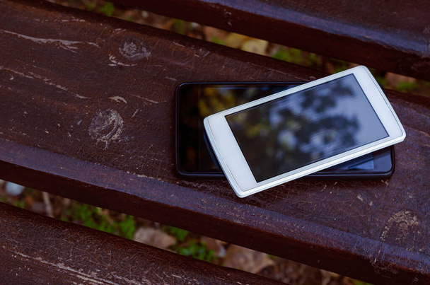 愛の概念日付電話やソーシャルネットワークなし.2台のスマートフォンが公園のベンチにある。茶色の木製のベンチに白と黒のデバイス。誰もいない - 写真・画像