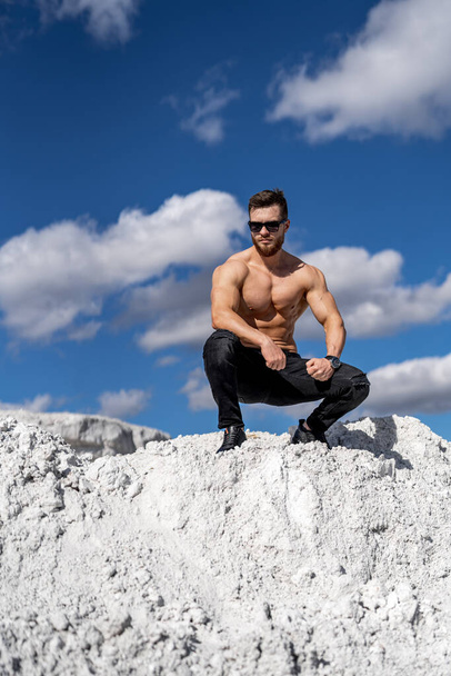 Man with perfect body pumping up muscles. Sitting in a quarry. Grey rocks background. - Foto, Bild