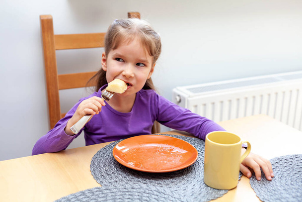 Young happy primary school european girl eating her food by herself, grabbing a last bite on a fork, empty plate and a mug before her. Children eating, self-reliant kids home activities, family meal - Photo, Image