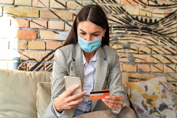 Pretty young woman with protective face mask using mobile phone anc credit card in the cafe - Фото, изображение