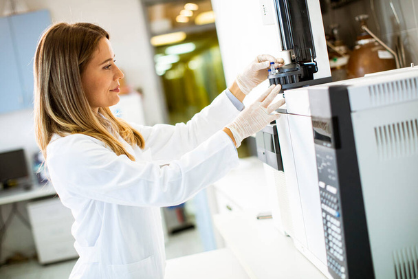 Mladá vědkyně v bílém laboratorním plášti dává lahvičku se vzorkem pro analýzu plynového chromatografu v biomedicínské laboratoři - Fotografie, Obrázek