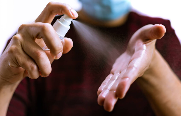 In a close-up view, you can see the hands of a Caucasian man receiving a spray of disinfectant liquid on his hands to disinfect them. In the background and out of focus, you can see that this man is wearing a blue surgical mask and a red sweater, all - Photo, image