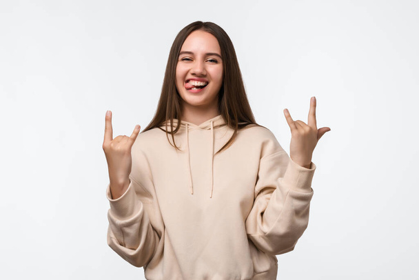 Emotiver Νεαρή γυναίκα με μακριά καστανά μαλλιά ίσια μαλλιά κάνει ROCK N ROLL υπογράψει, λέει: Θα ροκ αυτό το κόμμα, φορώντας κομψό μπεζ hoodie, αισθάνεται αυτοπεποίθηση. Αντίληψη ανθρώπινων συναισθημάτων - Φωτογραφία, εικόνα