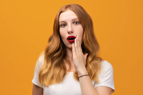 Pretty girl with wavy redhead, wearing white t-shirt expressing emotion of surprise standing isolated over yellow background - Foto, Imagem
