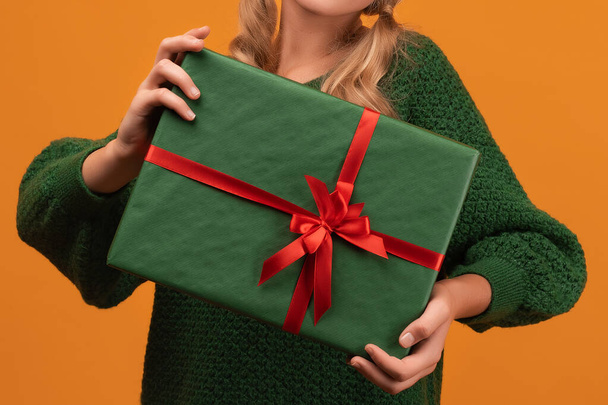 Image of Cropped view of girl in warm green sweater holding gift with red ribbon. Studio shot, yellow background, isolated. New Year Women's Day Birthday Holiday concept - Photo, Image