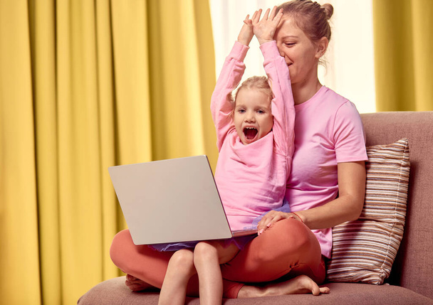 Anne ve kızı evde dizüstü bilgisayar kullanıyor. Pembe elbiselerle kanepede oturuyor. - Fotoğraf, Görsel