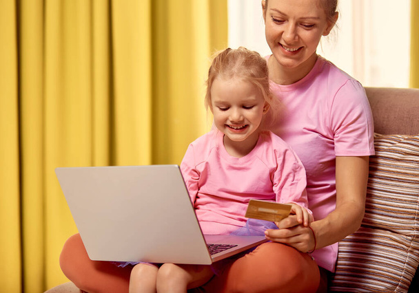 Achats ou paiements en ligne concept. Mère enseigne à sa fille de 4-5 ans en utilisant un ordinateur portable et une carte de crédit à la maison. Assis sur le canapé dans des vêtements roses - Photo, image
