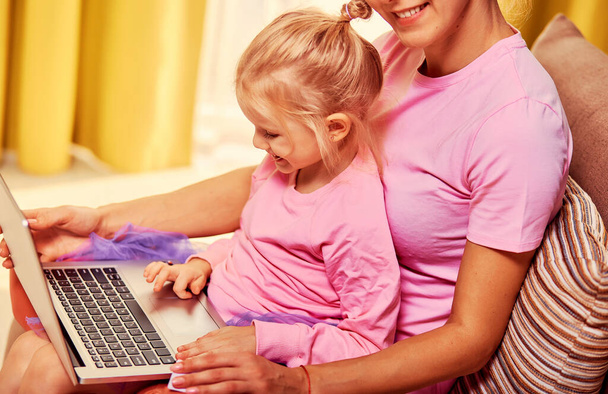 Online nakupování nebo platby koncept. Matka učí jeho 4-5 letou dceru používat notebook a kreditní kartu doma. Sedí na gauči v růžových šatech - Fotografie, Obrázek