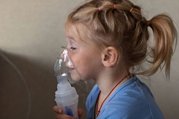 La niña hace inhalación con un nebulizador en casa, sentada en una silla. Aparato portátil para la obstrucción, asma bronquial. - Foto, imagen