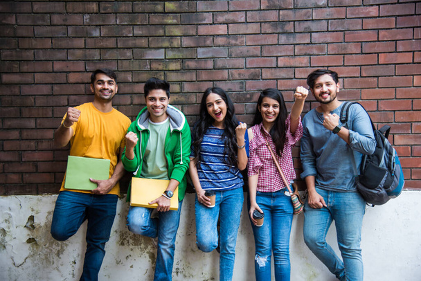 Wesoła indyjska azjatycka młoda grupa studentów lub przyjaciół śmiejących się razem siedząc, stojąc lub spacerując po kampusie - Zdjęcie, obraz