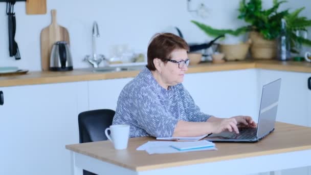 Idősebb nő, aki laptopot használ a webszörfözéshez a konyhájában. Az időskori foglalkoztatás, a társadalombiztosítás fogalma. Idősebb hölgy ül a munkahelyén, és jegyzetfüzetet gépel egy otthoni irodában. Fényképezte: - Felvétel, videó