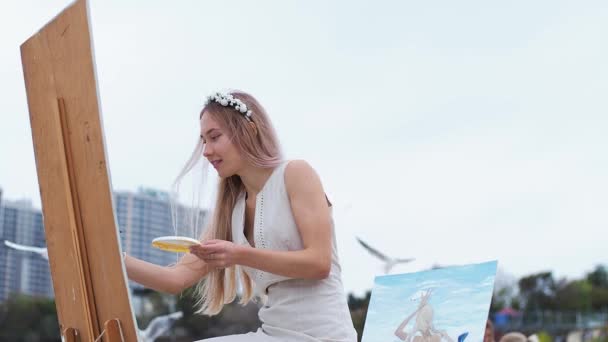 Jeune femme blonde peint le tableau sur la plage En plein air Sur fond de mouettes volantes - Séquence, vidéo