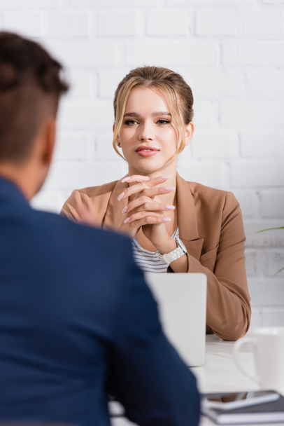 Mujer de negocios positiva mirando a su compañero de trabajo, mientras está sentado en el lugar de trabajo en primer plano borrosa - Foto, Imagen