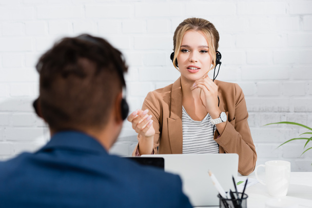 Kobieta konsultant w słuchawkach gestykulacji, siedząc w miejscu pracy na niewyraźnym pierwszym planie - Zdjęcie, obraz