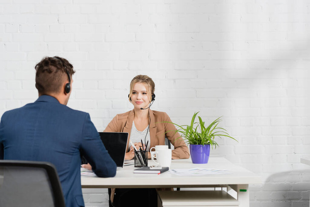 Pozitív női operátor fülhallgatóban nézi kollégáját, miközben a munkahelyén ül digitális eszközökkel - Fotó, kép