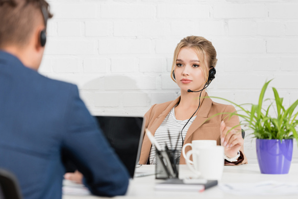 Mulher séria no fone de ouvido olhando para colega de trabalho, enquanto sentada no local de trabalho no escritório em primeiro plano turvo - Foto, Imagem