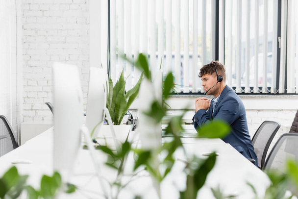 前景にぼやけた植物とオフィスの職場に座ってヘッドセットで深刻な演算子 - 写真・画像