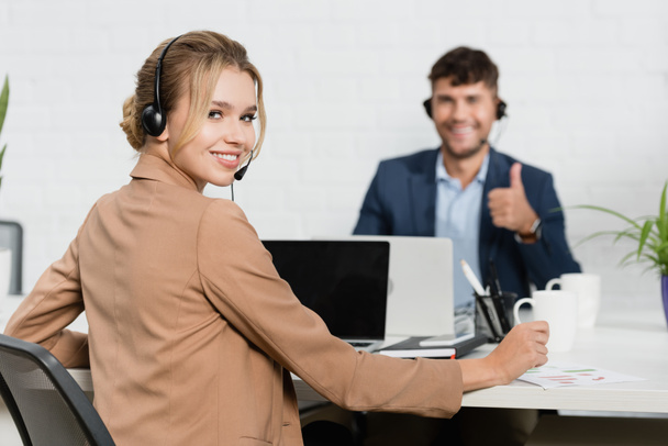 glimlachende vrouw in headset kijken naar camera, terwijl zitten op de werkplek met wazig collega op de achtergrond - Foto, afbeelding