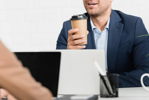 Vista recortada del trabajador de oficina con taza de papel hablando, mientras está sentado en la mesa con computadoras portátiles en primer plano borroso - Foto, Imagen