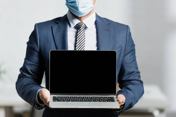 Vista recortada del ejecutivo en máscara médica, que muestra el ordenador portátil con pantalla en blanco sobre fondo borroso - Foto, imagen