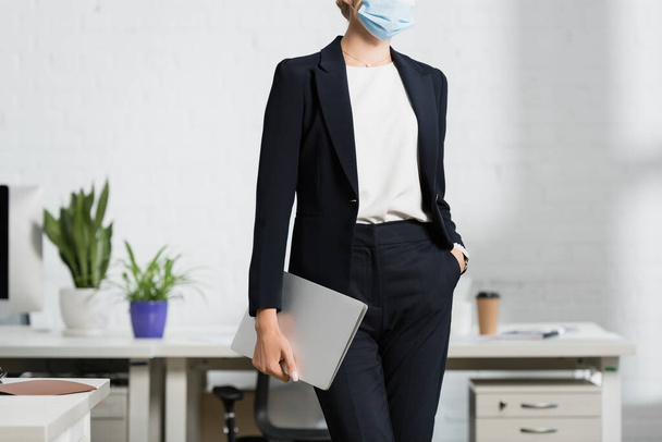 Cropped view of businesswoman with hand in pocket holding laptop, while standing in office on blurred background - Foto, imagen