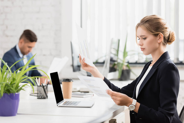 Εστιασμένη επιχειρηματίας που κοιτάζει γραφήματα σε φύλλα χαρτιού, ενώ κάθεται στο χώρο εργασίας σε θολή φόντο - Φωτογραφία, εικόνα