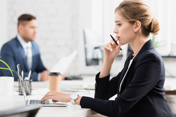 Zijaanzicht van attente zakenvrouw met pen typen op laptop, terwijl zitten op de werkplek op wazige achtergrond - Foto, afbeelding