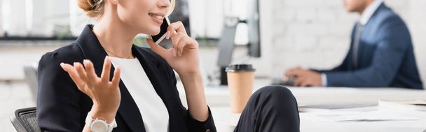 Vista recortada de una mujer de negocios sonriente haciendo un gesto, mientras habla por teléfono móvil en el lugar de trabajo sobre un fondo borroso, pancarta - Foto, Imagen