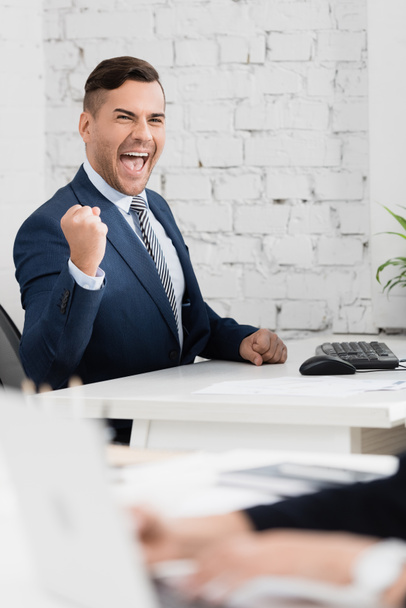 Excited businessman with yes gesture looking at camera, while sitting at table in office on blurred foreground - Photo, Image