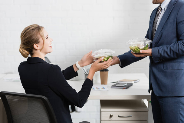 Happy businesswoman taking plastic bowl with meal from colleague, sitting at workplace - Photo, Image