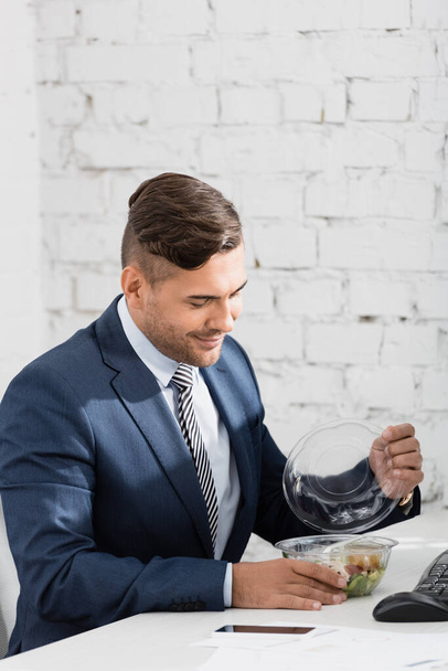 Homme d'affaires souriant ouvrant bol en plastique avec repas, tout en étant assis sur le lieu de travail - Photo, image