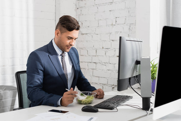 Empresario comiendo comida de un tazón de plástico, mientras está sentado en el lugar de trabajo con dispositivos digitales - Foto, Imagen