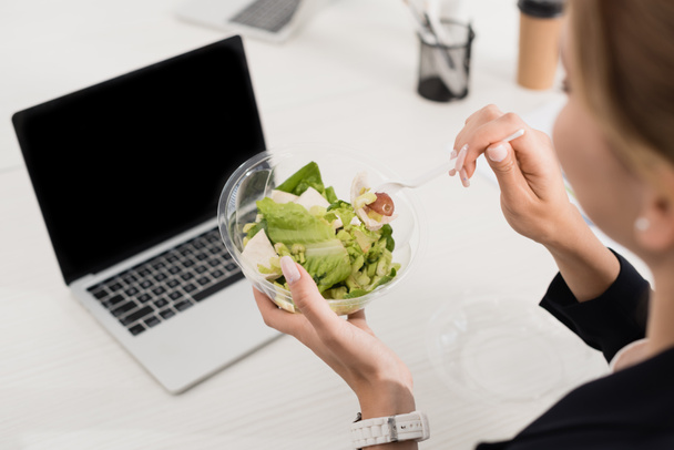 High angle view of businesswoman holding plastic fork and meal in bowl at workplace on blurred foreground - Фото, изображение