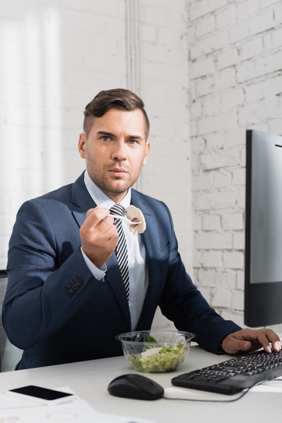 Businessman eating meal from plastic bowl, while looking at camera at workplace with digital devices - Photo, image