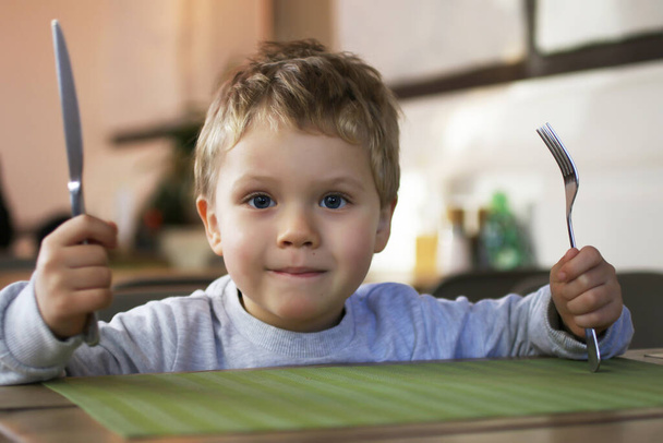 Ребенок показывает, что хочет есть. Парень держит вилку и нож над столом. Интерьер в современном ресторане. - Фото, изображение