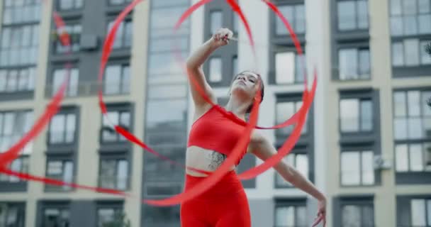 Jonge vrouw in het rood voert callisthenics met lint in de libing appartement werf, gymnast doet acrobatische oefeningen in een residentiële ruimte, fitness in de stedelijke omgeving, 4k 120p Prores HQ - Video