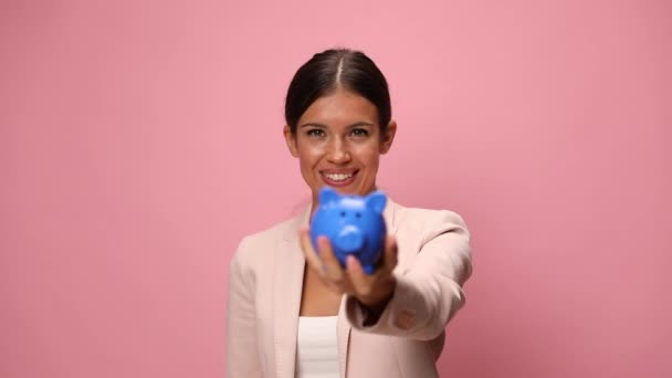 usmívající se mladá žena v růžovém obleku dávat mince v prasátko banky, přikyvování, dělat palce nahoru gesto na růžovém pozadí - Záběry, video