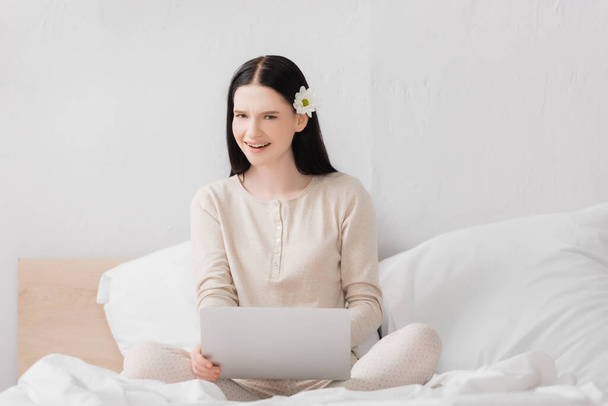 smiling freelancer with vitiligo and flower in hair using laptop in bedroom  - Photo, Image