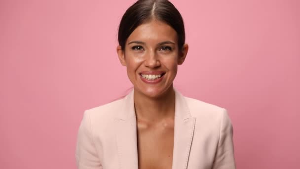 mosolygós fiatal üzletasszony rózsaszín öltöny mögött bujkál malac bank és tartsa karját a levegőben ünneplő rózsaszín háttér - Felvétel, videó