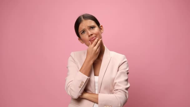 confused young businesswoman in pink suit crossing arms, touching chin and thinking, making uncertain faces, grimacing and desperately holding hands on temples gesturing on pink background - Footage, Video