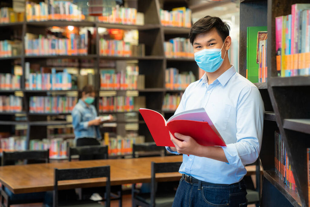 Dwóch studentów azjatyckich uniwersytetów noszących maski twarzy i stojących w bibliotece społecznej odległości od innych 6 stóp, aby uniknąć rozprzestrzeniania się koronawirusu na Uniwersytecie. - Zdjęcie, obraz