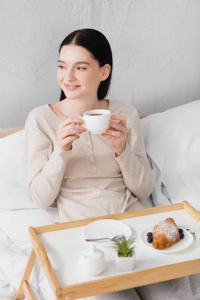 young happy woman with vitiligo holding cup of tea near breakfast on tray - Photo, Image