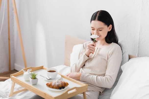 young brunette woman with vitiligo smelling flower near breakfast on tray - Photo, Image